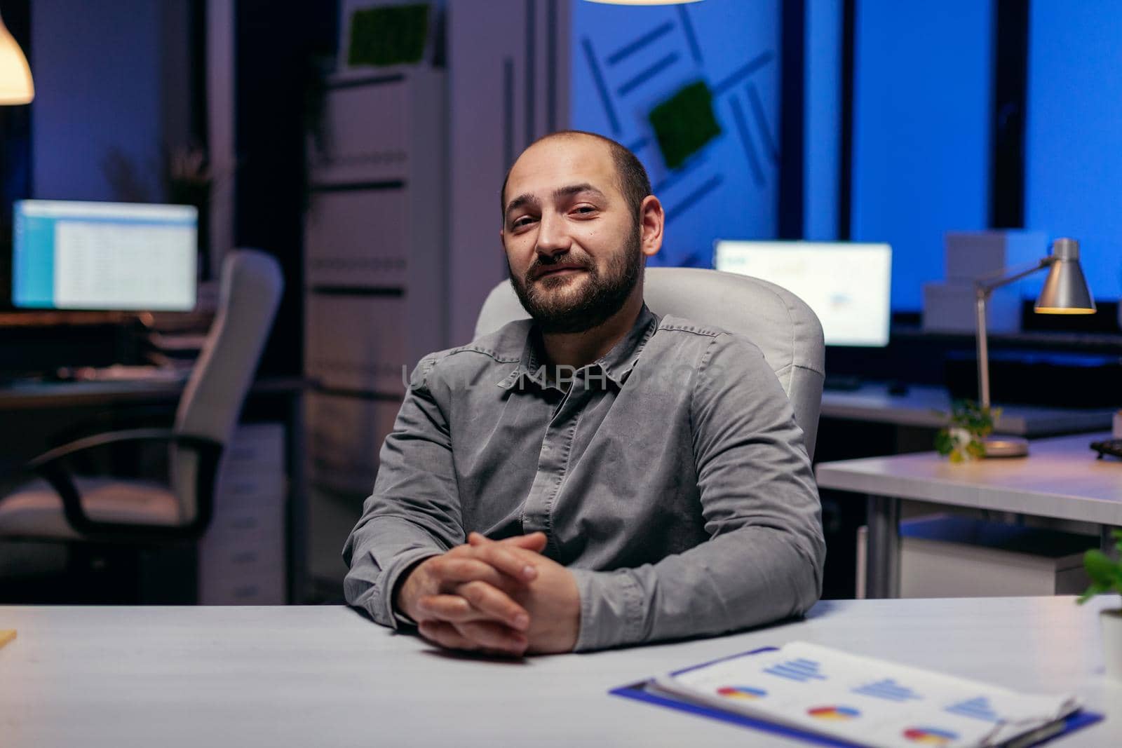 Portrait of concentrated manager smiling at camera thinking about success. Smart businessman sitting at his workplace in the course of late night hours doing his job.