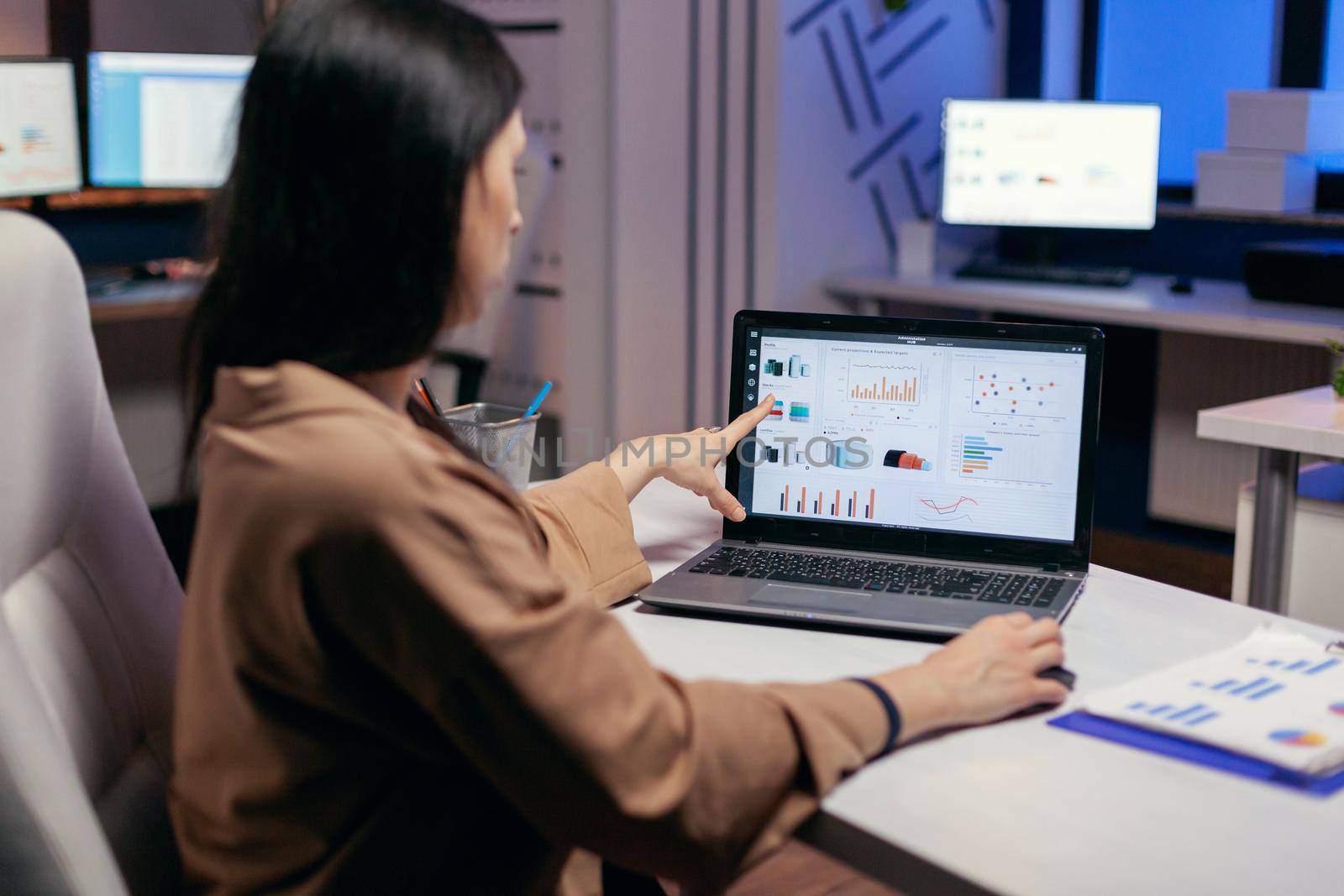 Busy employee pointing at graph working to finish a deadline wprlomg late. Businesswoman looking at statistics on laptpop screen sitting at her workplace doing overtime.