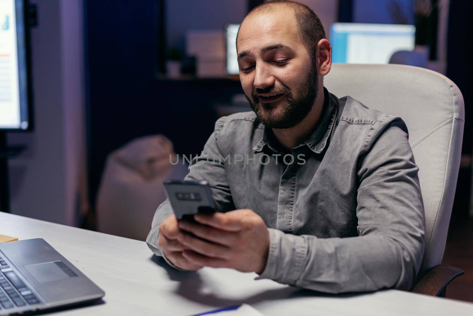 Entrepreneur writing text on smartphone sitting at desk in empy workplace. Businessman using his cell to text message while working late at night in the office to finish a deadline.