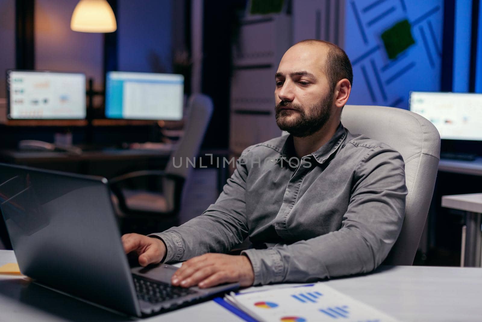 Caucasian entrepreneur working on deadline in dark office typing on laptop. Smart businessman sitting at his workplace in the course of late night hours doing his job.