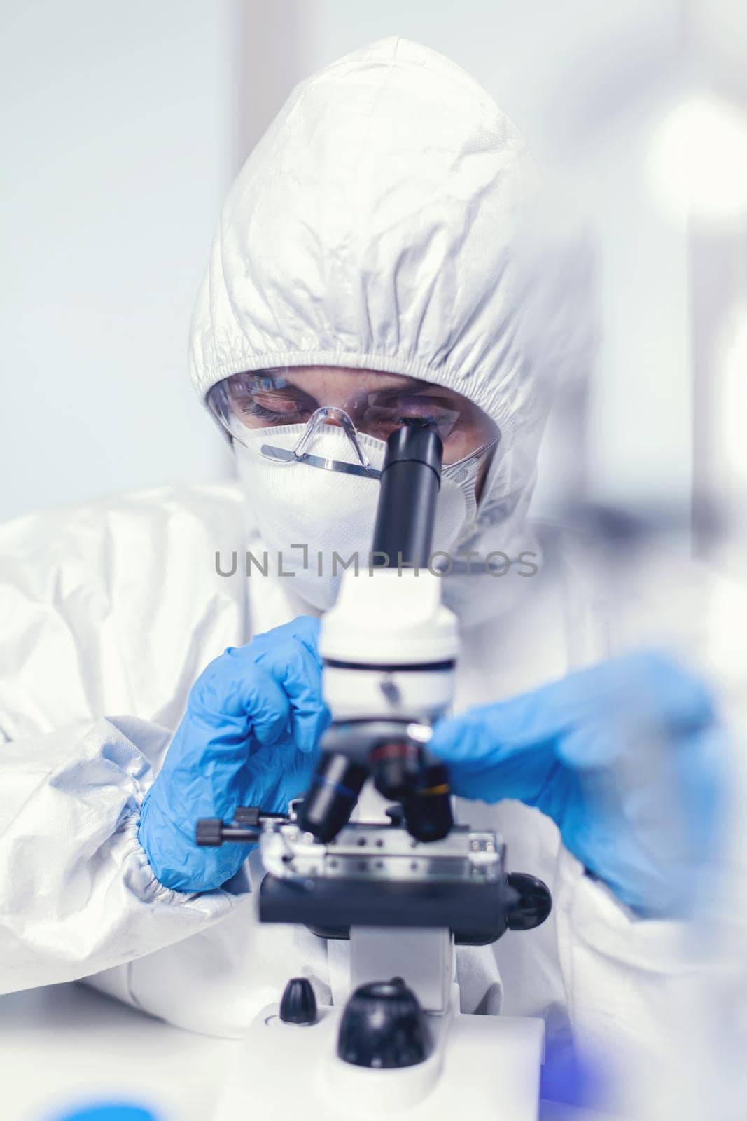 Medical engineer in ppe suit in modern laboratory looking at coronavirus sample using microscope. Chemist researcher during global pandemic with covid-19 checking sample in biochemistry lab