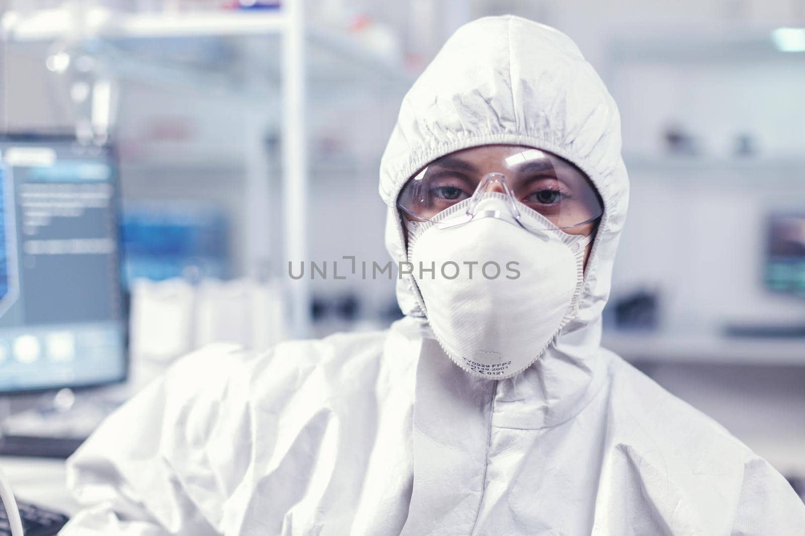 Portrait of exhausted chemist looking at camera wearing coverall in modern equipped lab. Tired woman scientist in biotechnology laboratory wearing protective suit during global epidemic.