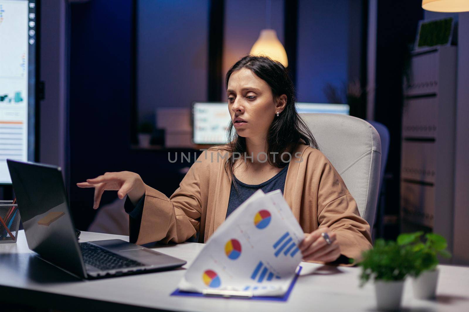 Tired employee reading article to help her finish deadline sitting at desk in empty office. Woman checking notes on clipboard while working on important project doing overtime.