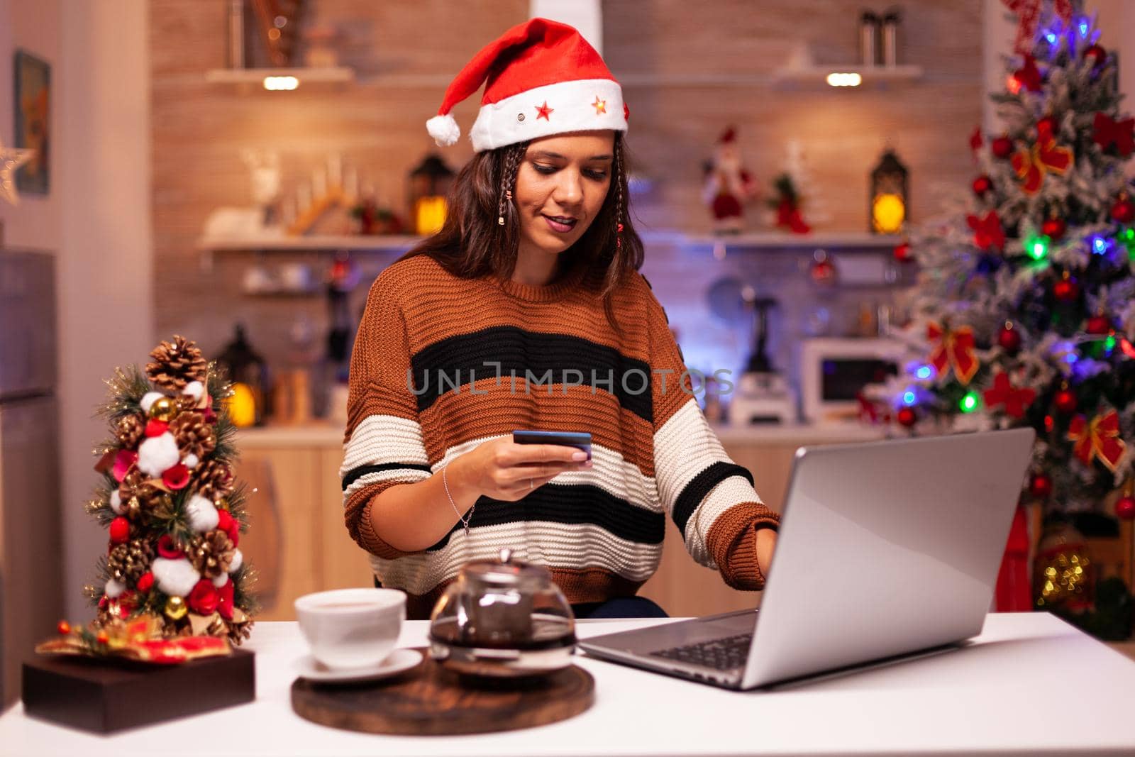 Traditional adult doing online christmas shopping for family gifts in kitchen with winter decorations, lights and ornaments for tree at home. Woman using credit card payment for purchase