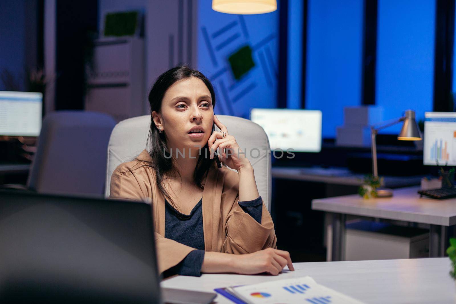 Successful businesswoman having a phone conversation in the evening sitting at workplace. Woman entrepreneur working late at night in corporate business doing overtime in the course of phone call.