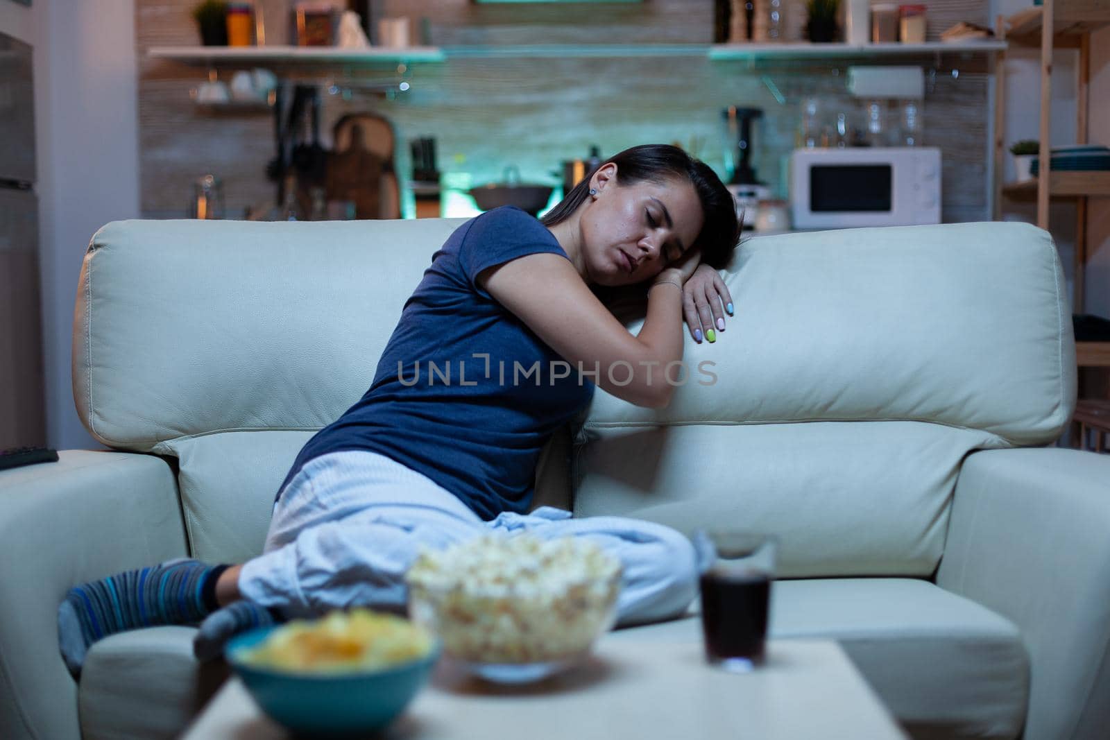 Portrait of woman sleeping on couch in living in front of television. Tired exhausted lonely sleepy lady in pajamas falling asleep sitting on cozy sofa, closing eyes while watching movie at night.