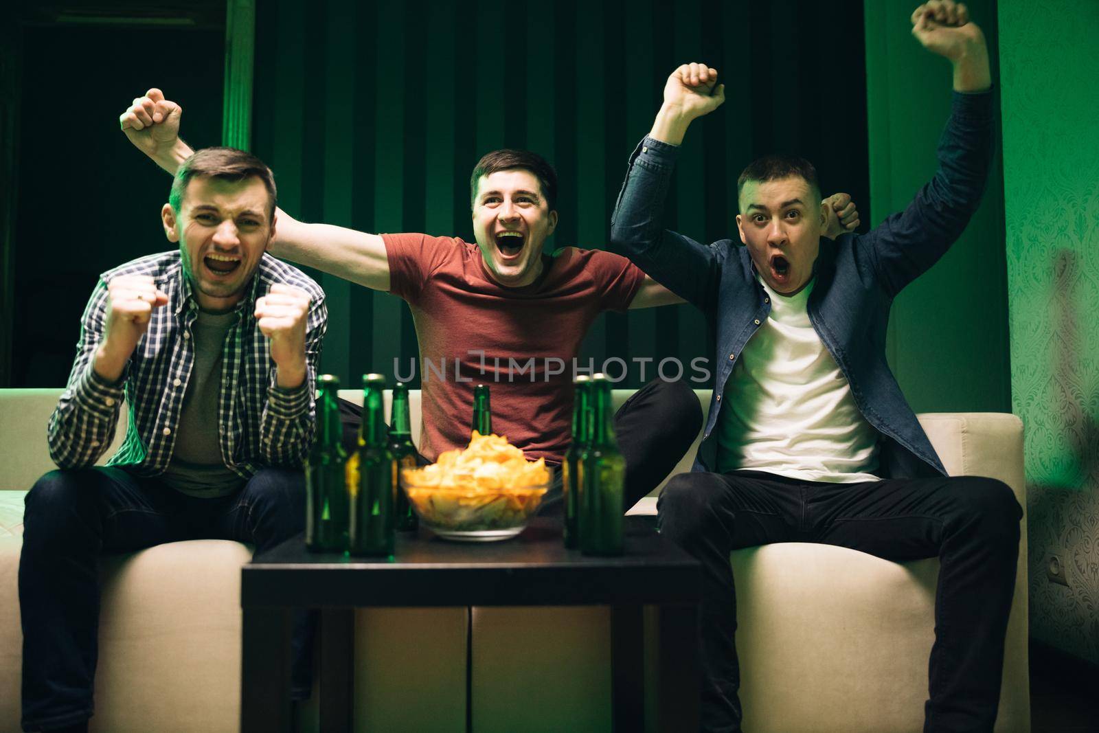Cheerful happy Caucasian guys cheering for favorite team. Loyal football fans supporting their team. Men watching TV with sport channel late at night and drinking beer.