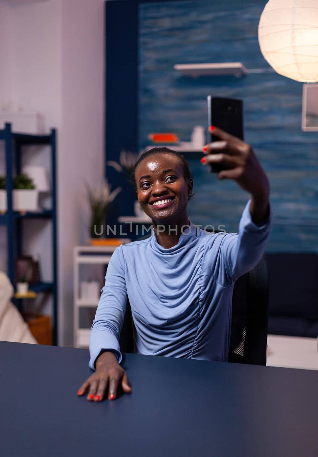 Cheerful dark skinned woman taking a self portrait using smartphone late at night from home. Busy focused freelancer using modern technology network wireless doing overtime.