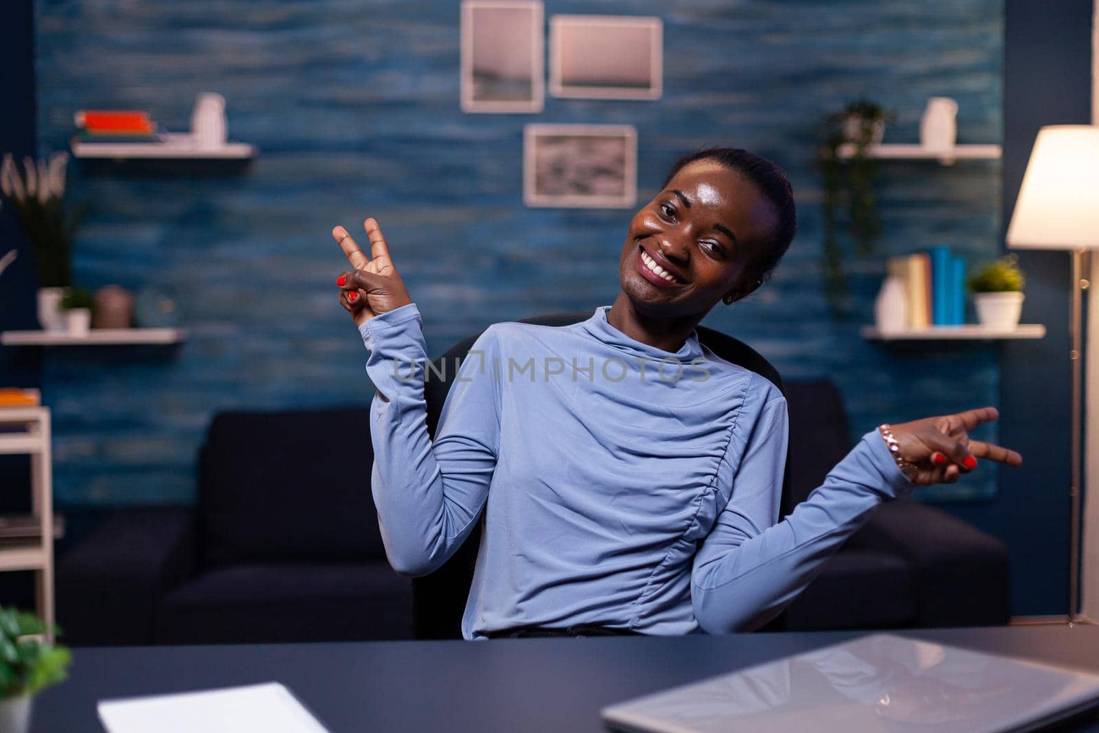 Cheerful dark skinned business woman showing victory sign looking at camera working late at night from home. . Black woman enjoy e-learning process, easy application usage overtime.
