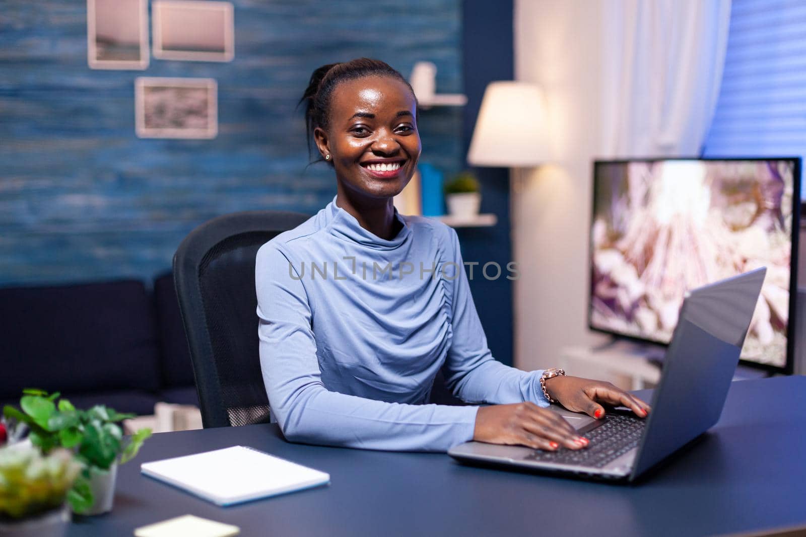 Smiling dark skinned woman smiling at camera sitting at desk working late at night from home office. Black freelancer working with remotely team chatting virtual online conference.