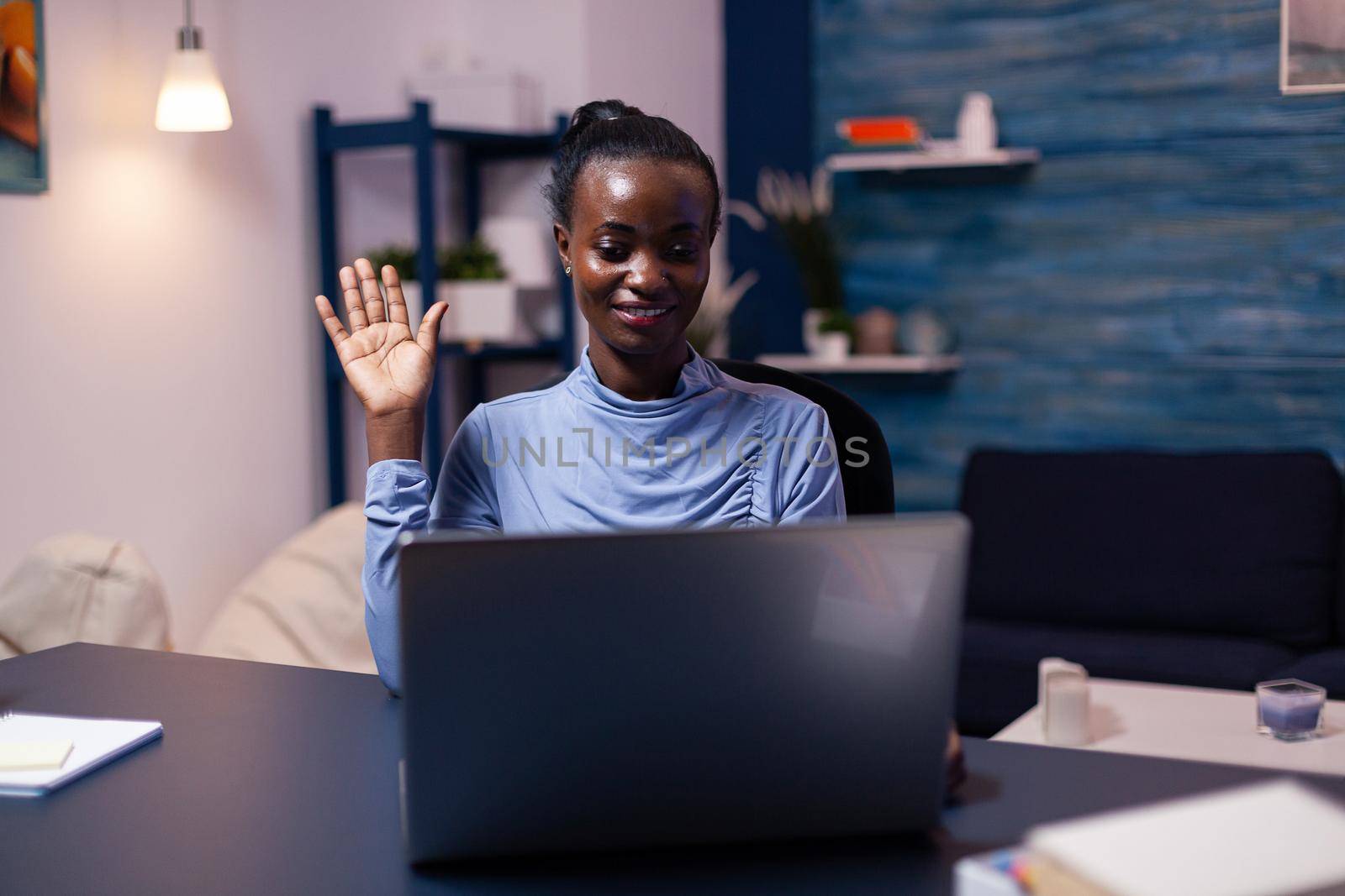 African businesswoman greeting people during video conference late at night in home office. Black freelancer working with remotely team chatting virtual online conference.