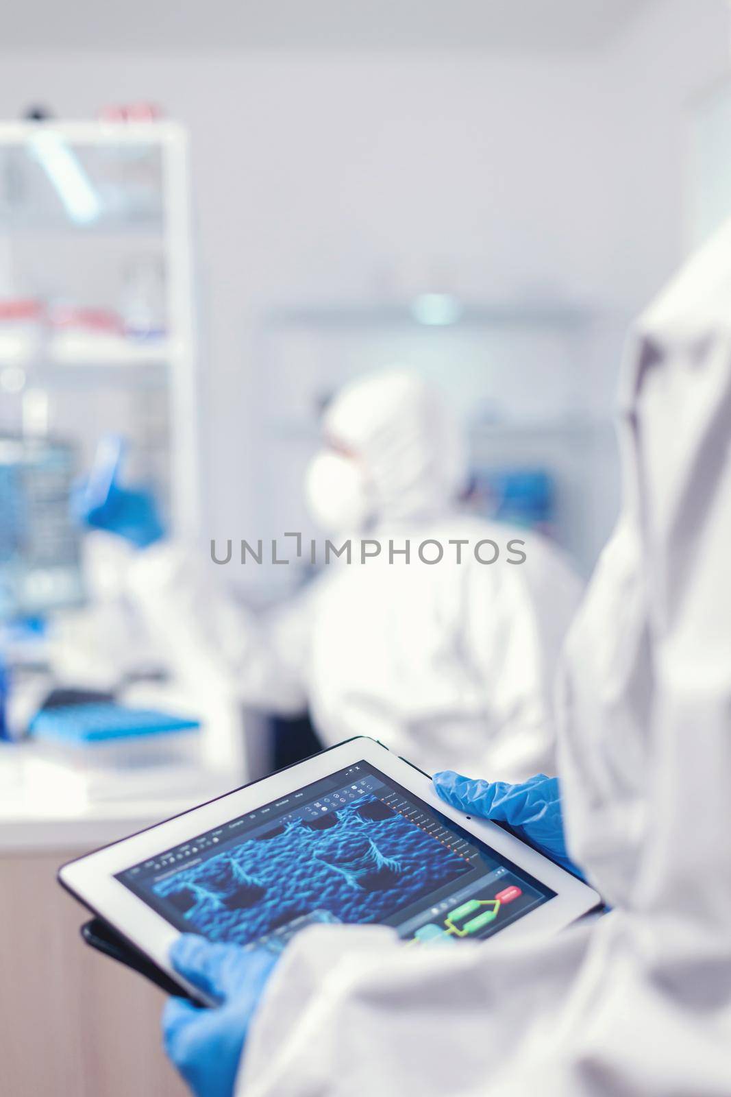 Chemist dressed in protection suit for coronavirus holding tablet pc in laboratory. Team of scientists conducting vaccine development using high tech technology for researching treatment against covid19