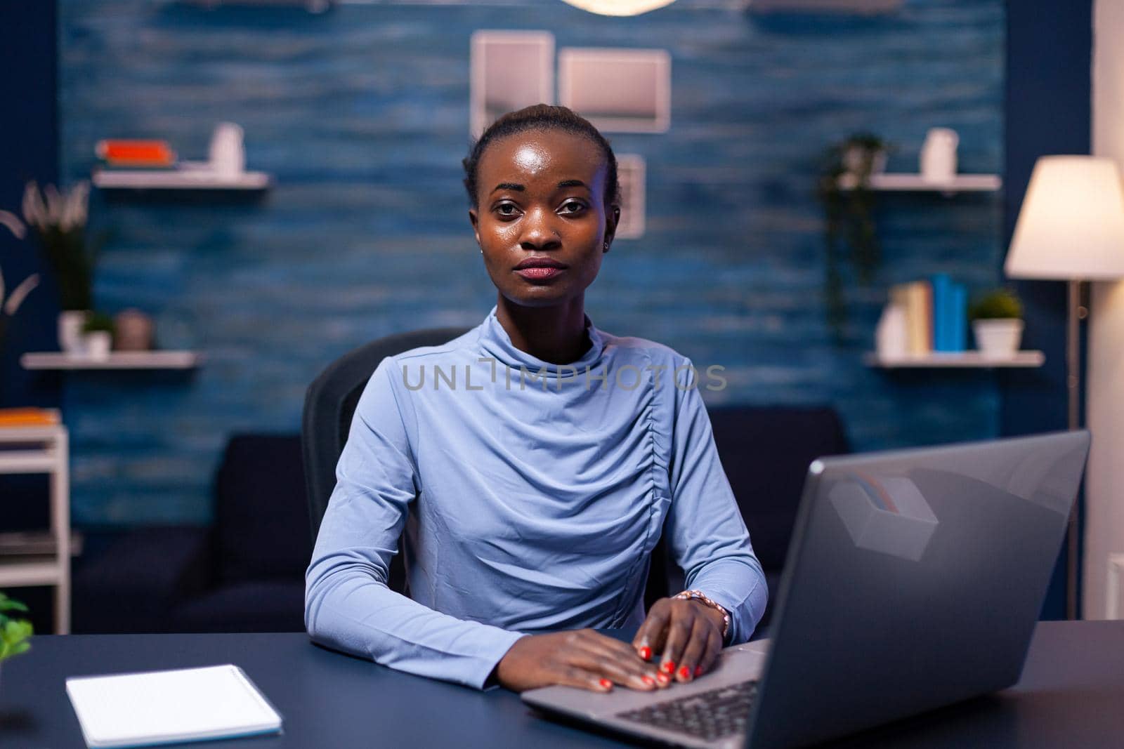 African business woman looking seriously at camera in the course of working on project. Black entrepreneur sitting in personal workplace writing on keyboard.