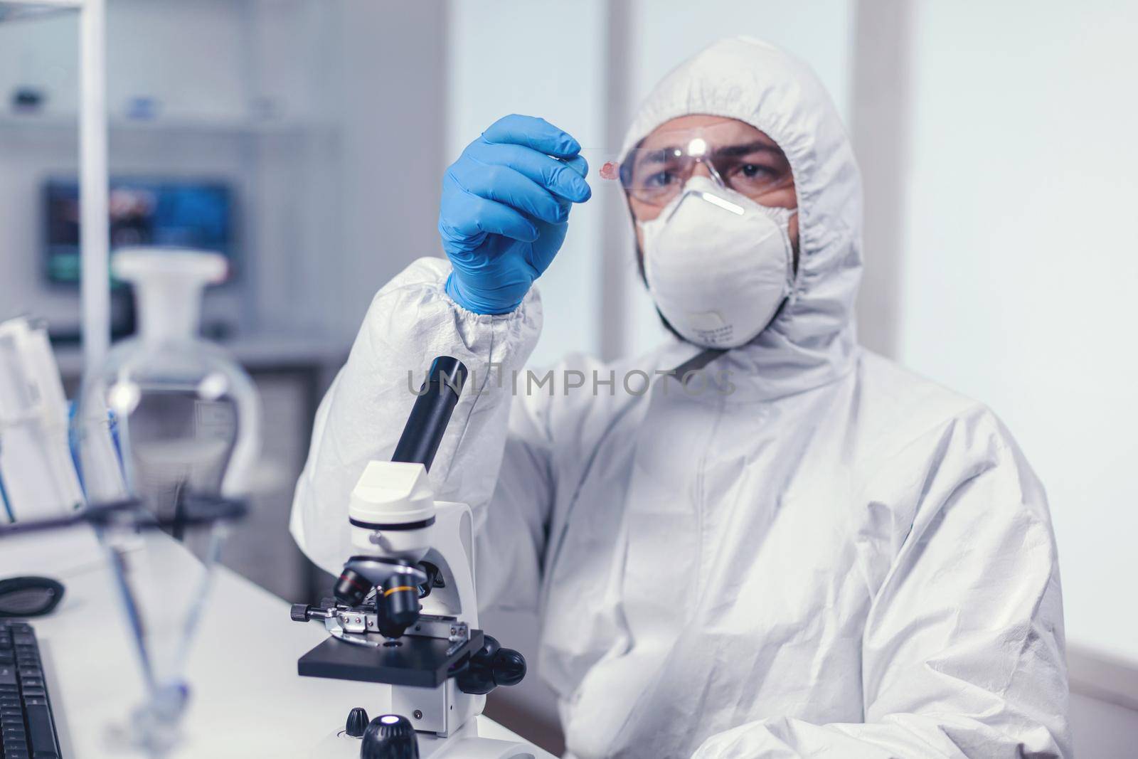Scientist looking at a microscope slide dressed in coverall during coronavirus. Examining vaccine evolution using high tech for researching treatment against covid19 virus.