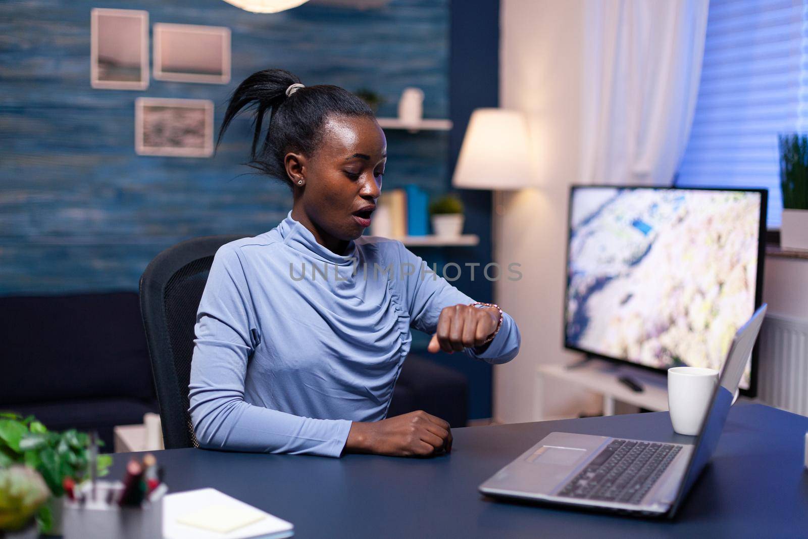 African business woman checking time looking at wrist watch working late on deadline from home office. Nervous black manager doing overtime working on computer.