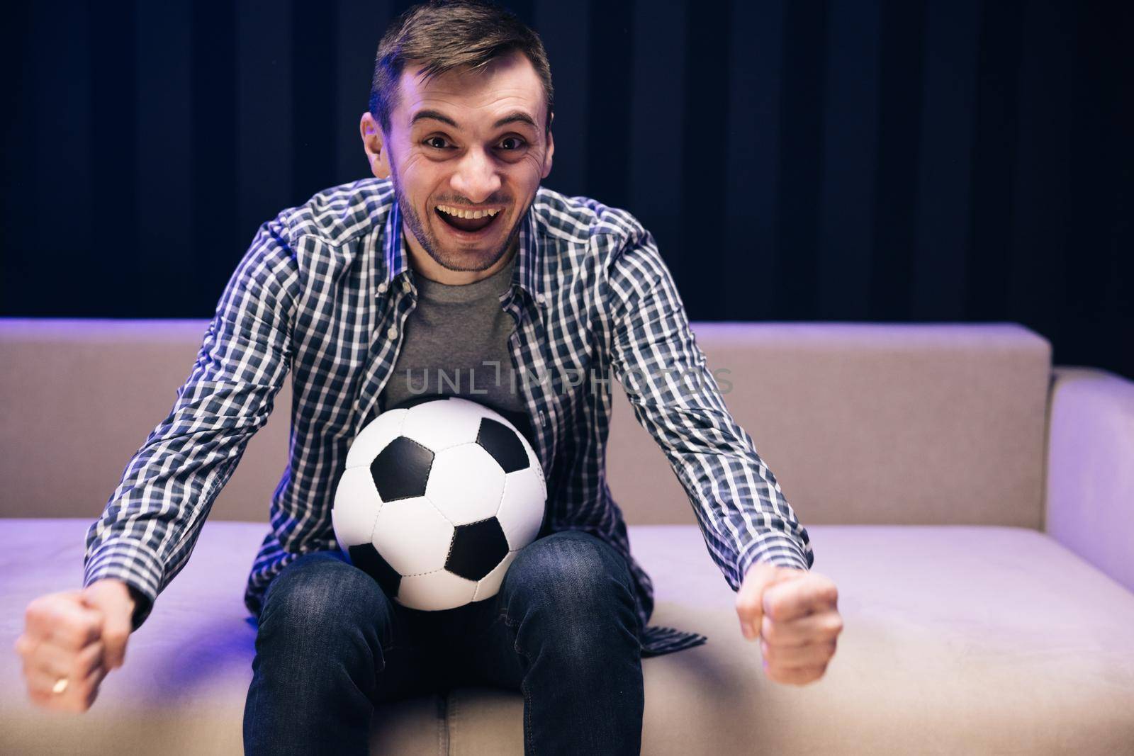 Sports Fan Reaction Concept. Fun guy 30s football fan cheer up support favorite team hold soccer ball in t-shirt in dark living room. People emotions sport leisure lifestyle.