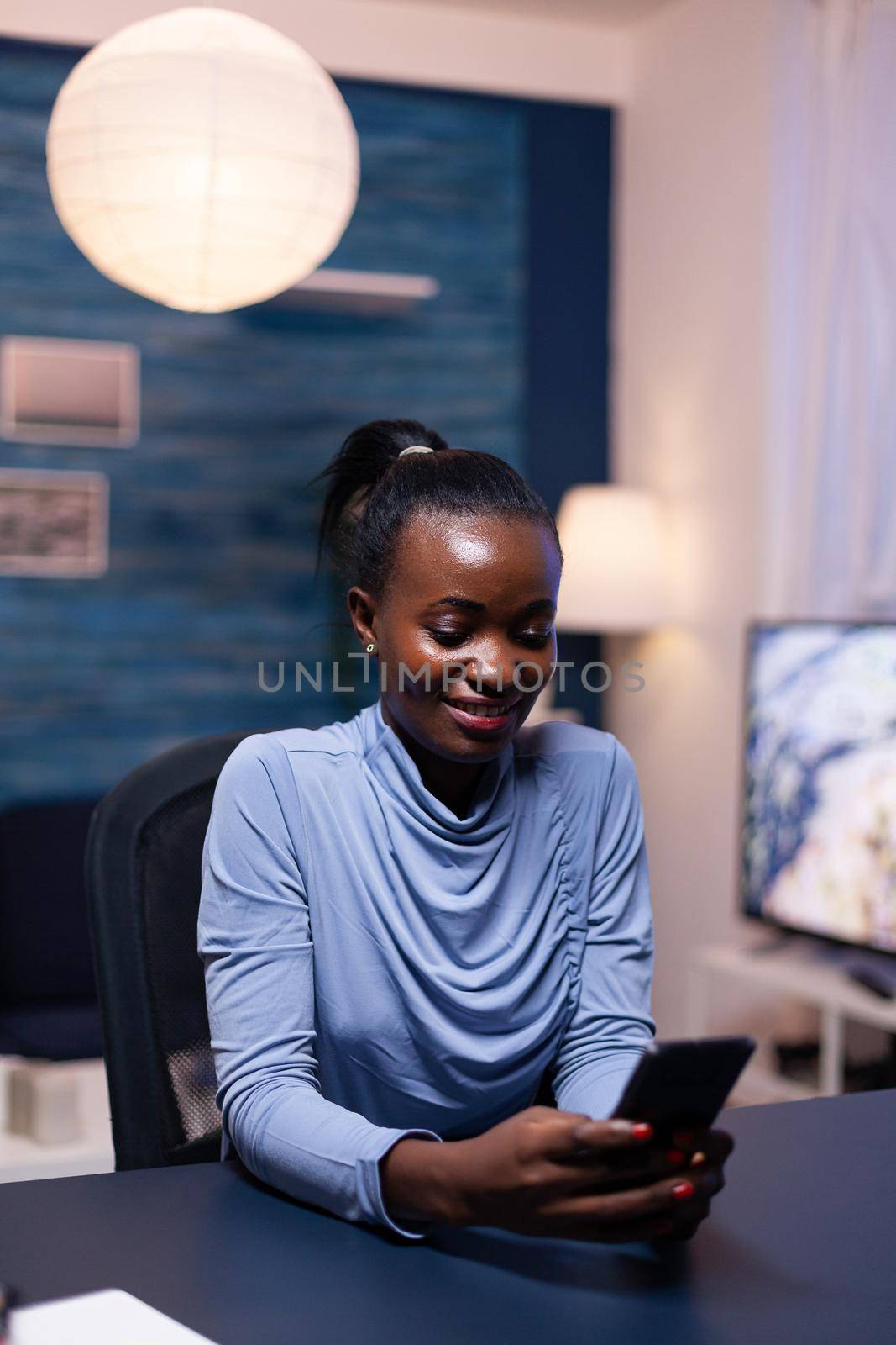 Cheerful african business woman texting on smartphone late at night in home office working remote. Employee using modern technology network wireless doing overtime.