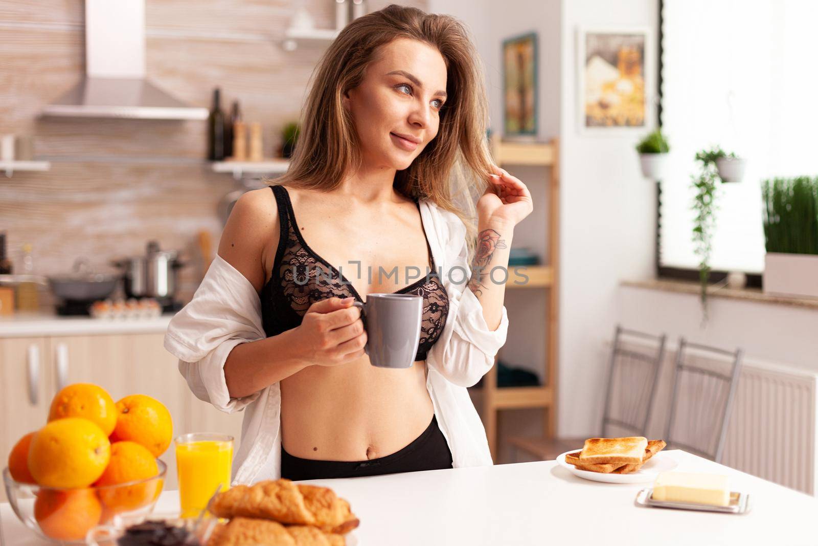 Young woman wearing sexy lingerie during breakfast in home kitchen. Attractive lady with tattoos in seductive underwear holding cup of tea relaxing in the kitchen smiling.