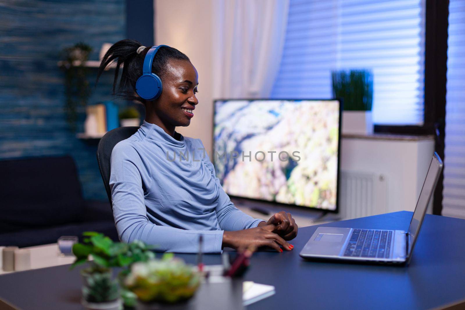 Cheerful black woman listening music wearing headset during work from home office sitting at desk looking at computer.. African freelancer creating new project working late.