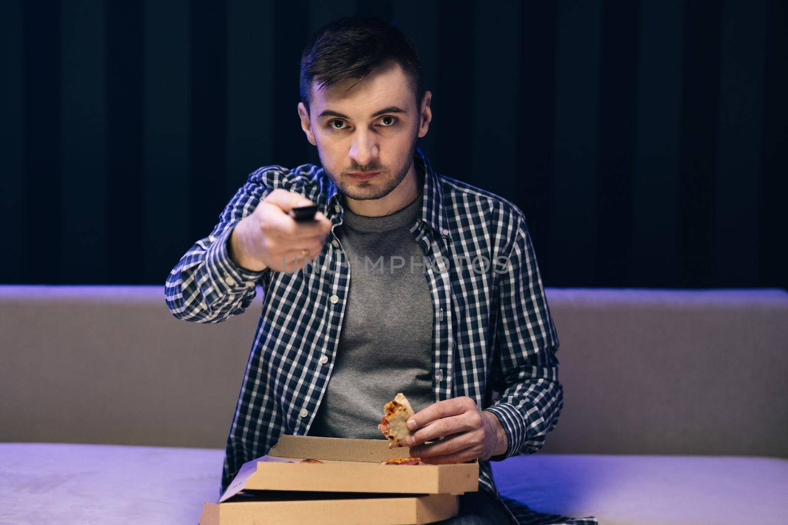 Portrait of young hipster man eating pizza, watching favorite film on tv while relaxing at sofa in the living room. Man eating Italian food. Relaxation cinema channels resting by uflypro