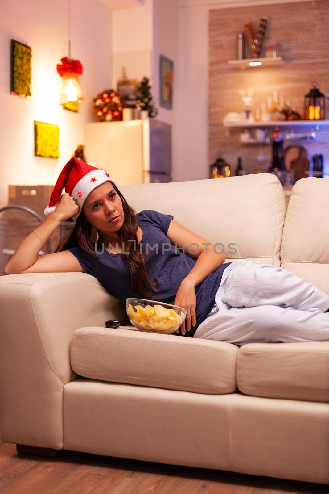 Caucasian female resting on sofa watching christmas entertainment film on television enjoying winter season in xmas decorated kitchen. Woman with red santa hat celebrating christmastime