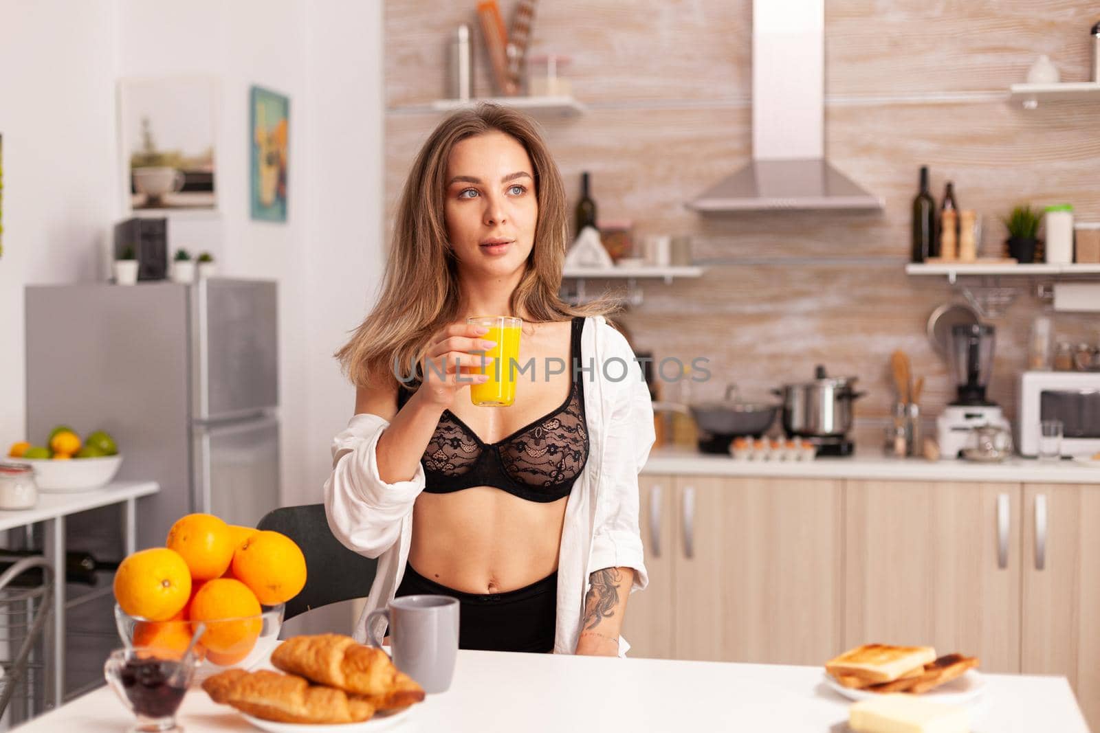 Woman holding glass with fresh orange juice during breakfast wearing sexy black lingerie. Young sexy seductive blode lady with tattoos drinking healthy, natural homemade orange juice, refreshing sunday morning