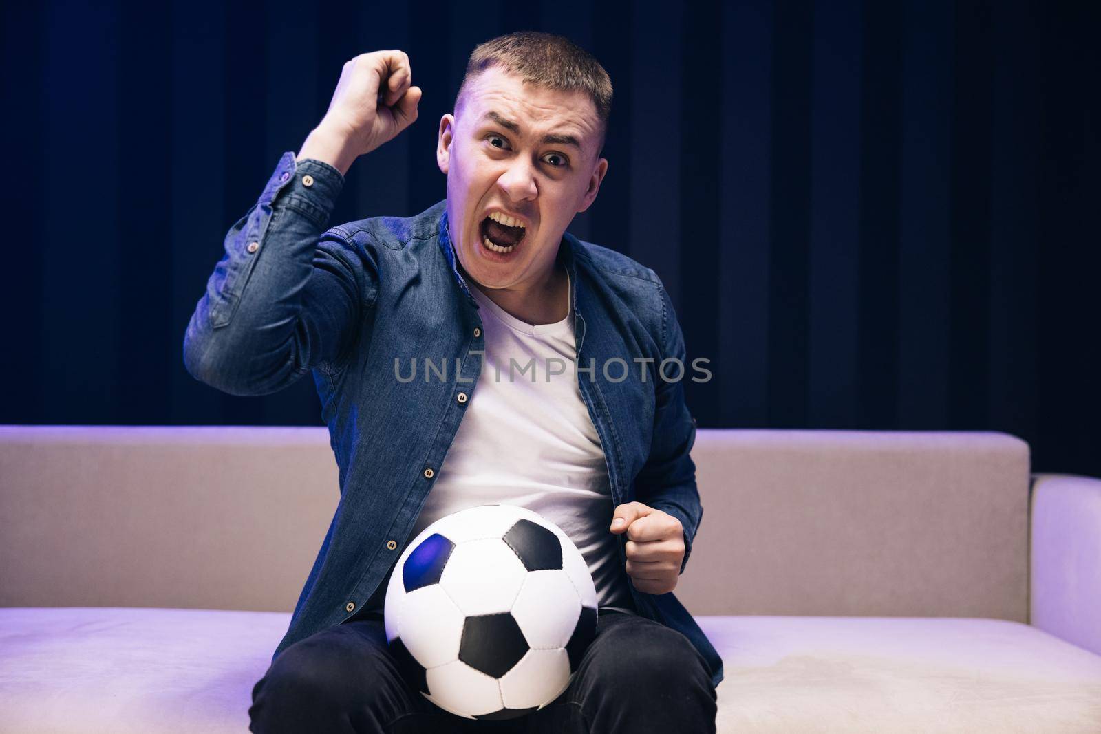 Young fun guy 25s football fan cheer up support favorite team hold ball in denim jacket white t-shirt in dark living room. People emotions sport leisure lifestyle concept.