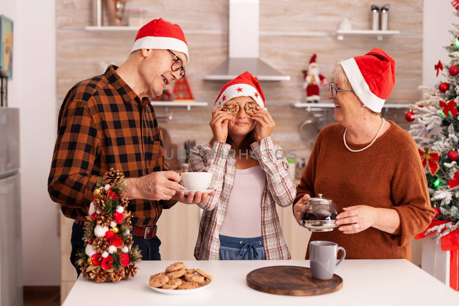 Happy family celebrating christmas holiday spending time together in xmas decorated kitchen enjoying winter season. Grandchild wearing santa hat having fun with delicious cookies during christmastime