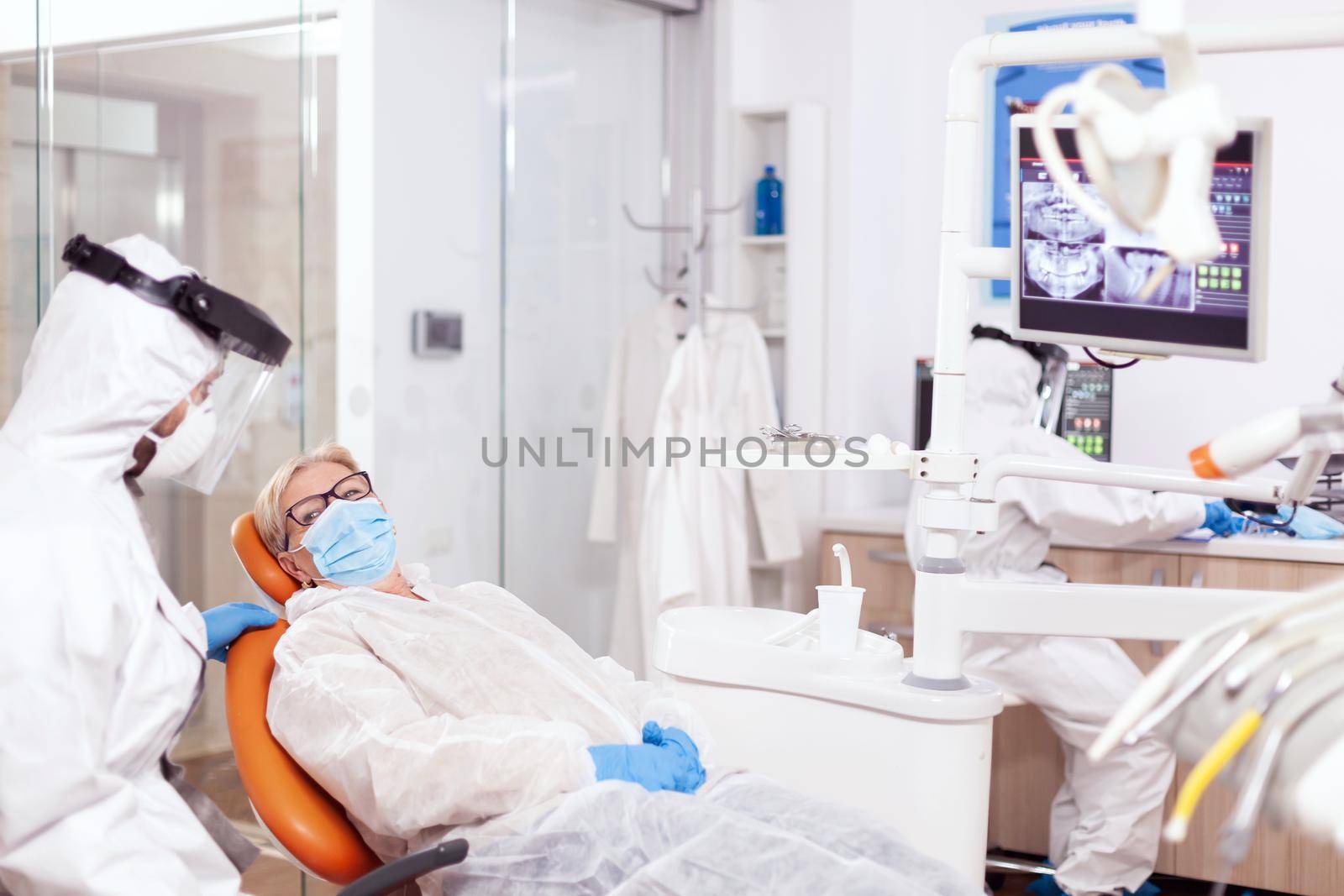 Dentist wearing protection agasint coronavirus during stomatology consultation of senior patient. Elderly woman in protective uniform during medical examination in dental clinic.