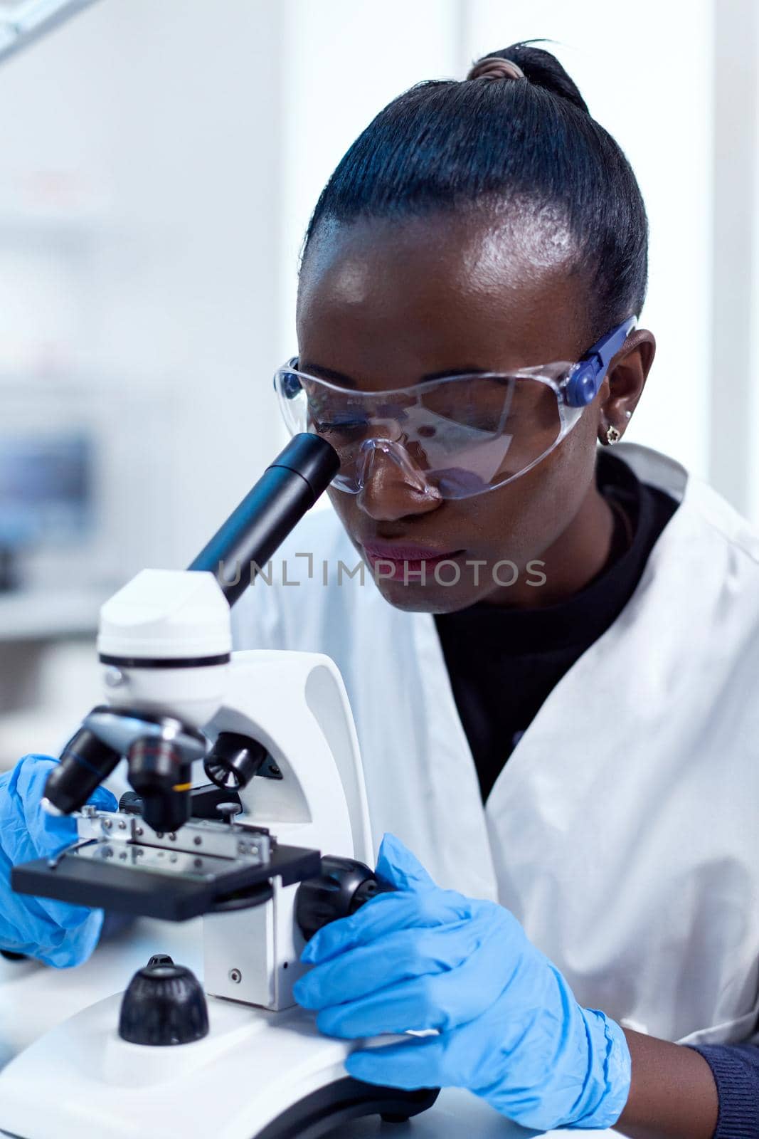 Close up of chemist of african ethnicity doing sample analysis looking through microscope. Black healthcare scientist in biochemistry laboratory wearing sterile equipment.