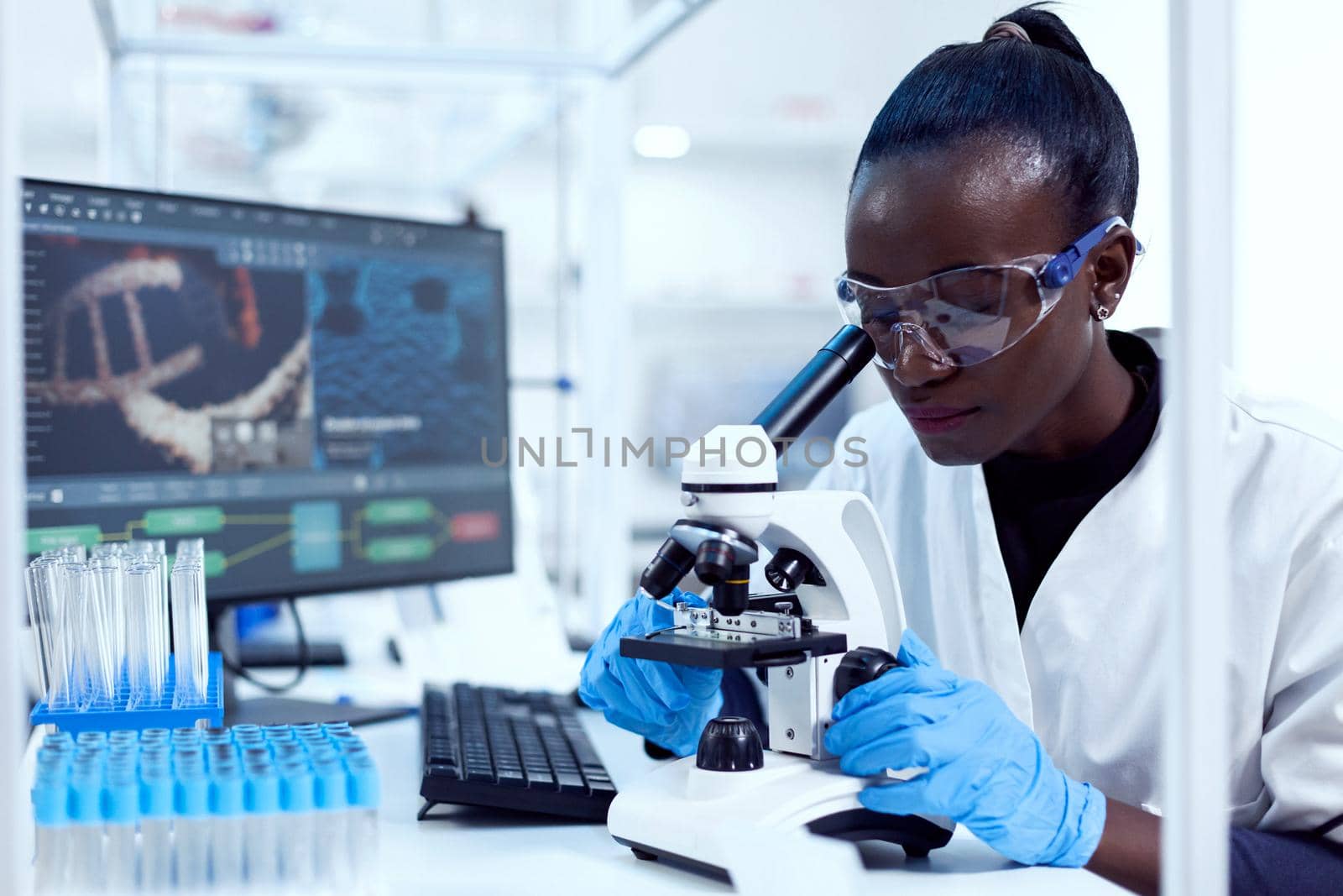 Virologist of african ethnicity sitting at her workplace neat test tubes looking through microscope. Black healthcare scientist in biochemistry laboratory wearing sterile equipment.