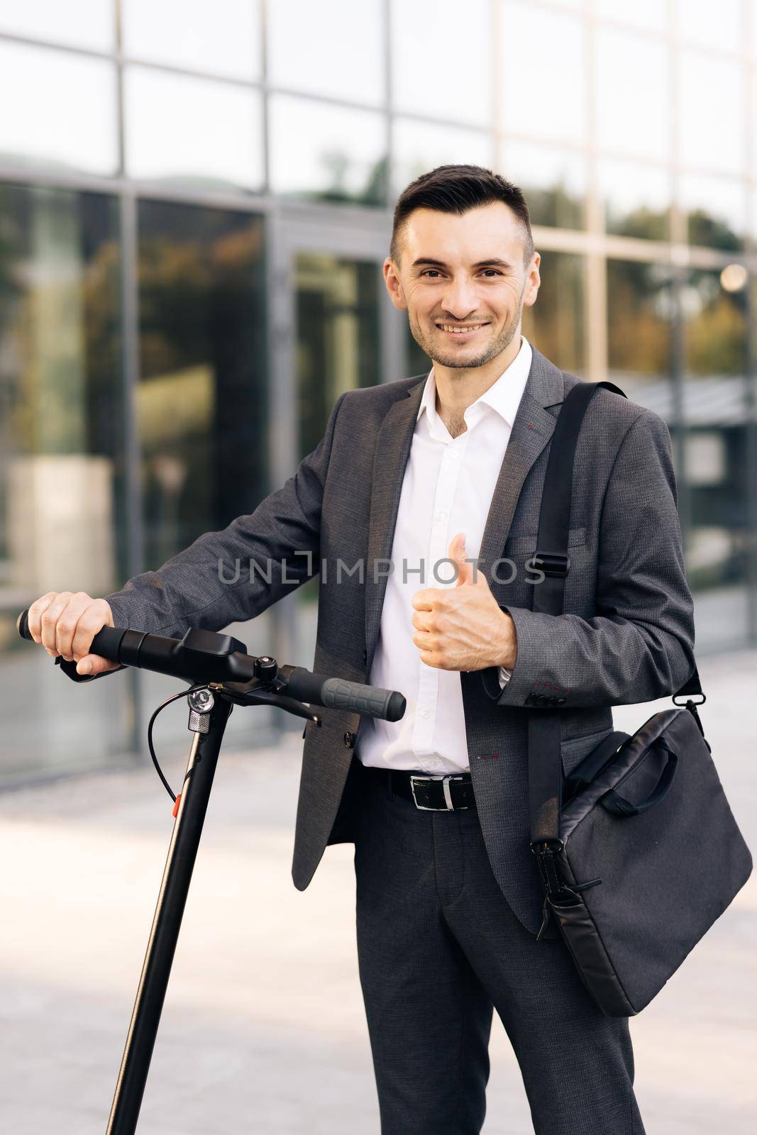 Handsome businessman with his electric scooter in front the business building. Young businessman in elegant suit standing outdoors, cheerfully smiling and showing thumb up. by uflypro