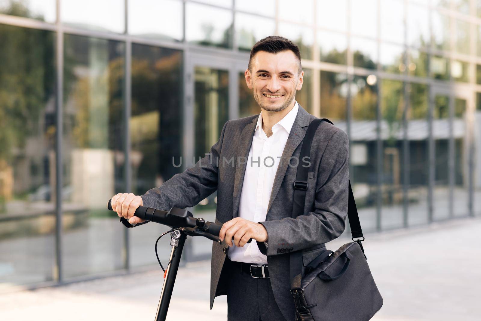 Confident businessman standing with electric scooter and looking camera. Happy successful businessman in a city. Ecology and urban lifestyle.
