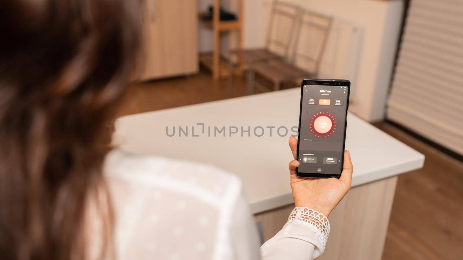 Woman in home kitchen using phone app to remotely control lighting system. Person in apartment holding telephone with touchscreen and app for lights.