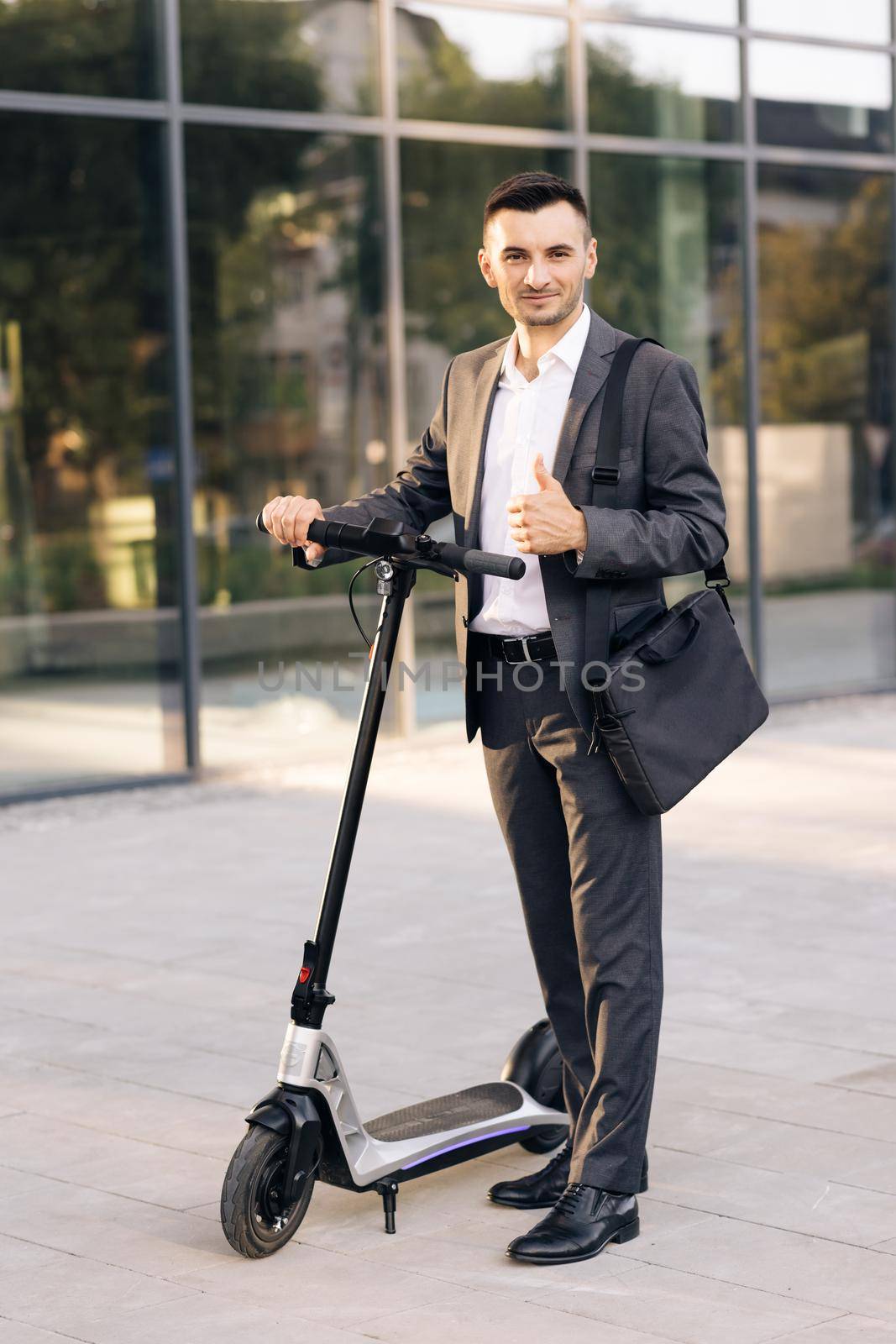 Young businessman in elegant suit standing outdoors, cheerfully smiling and showing thumb up. Handsome businessman with his electric scooter in front the business building by uflypro