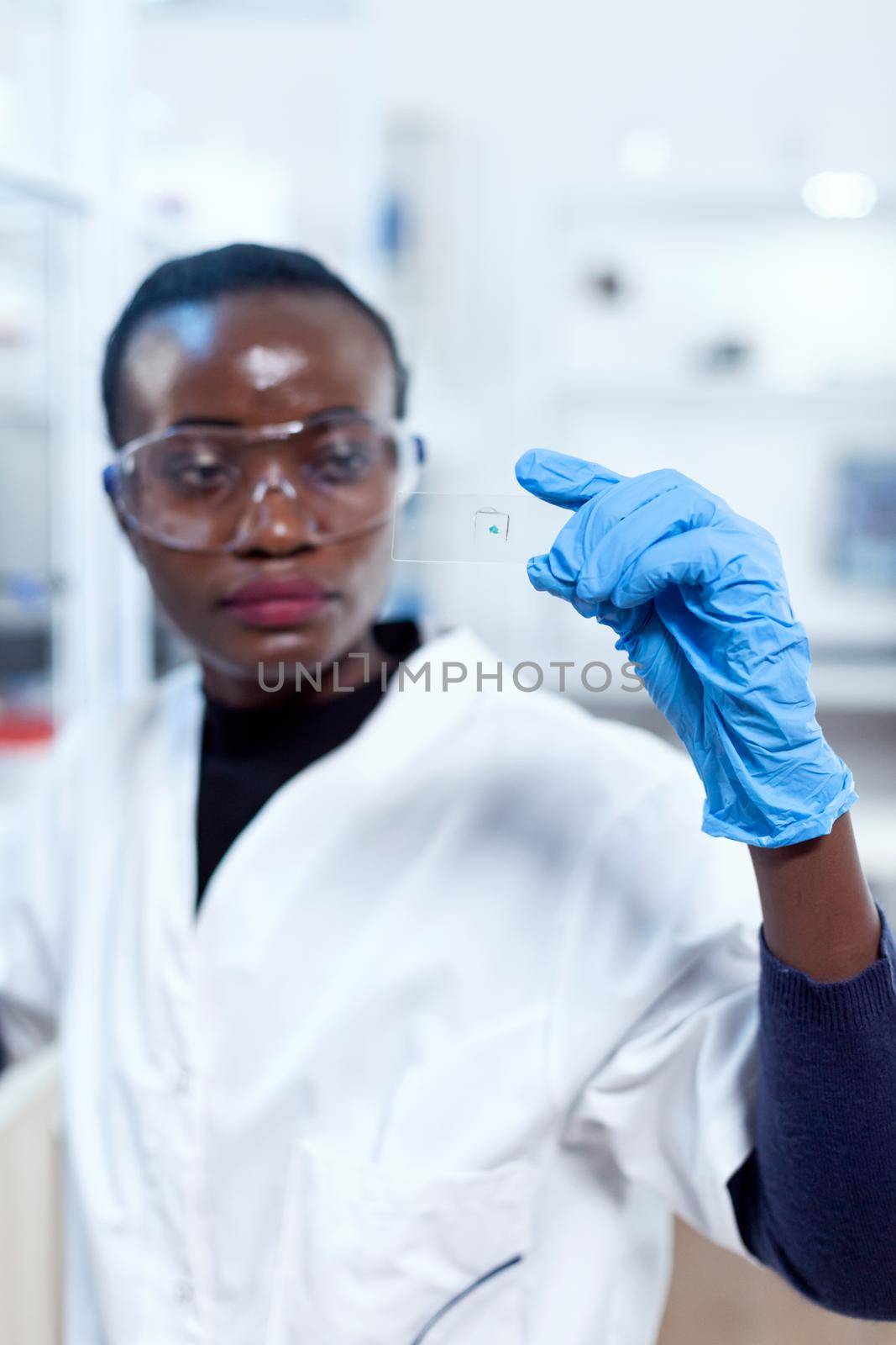 Close up of african biochemists holding microscope sample on glass slide with protective gloves. Black healthcare scientist in biochemistry laboratory wearing sterile equipment.