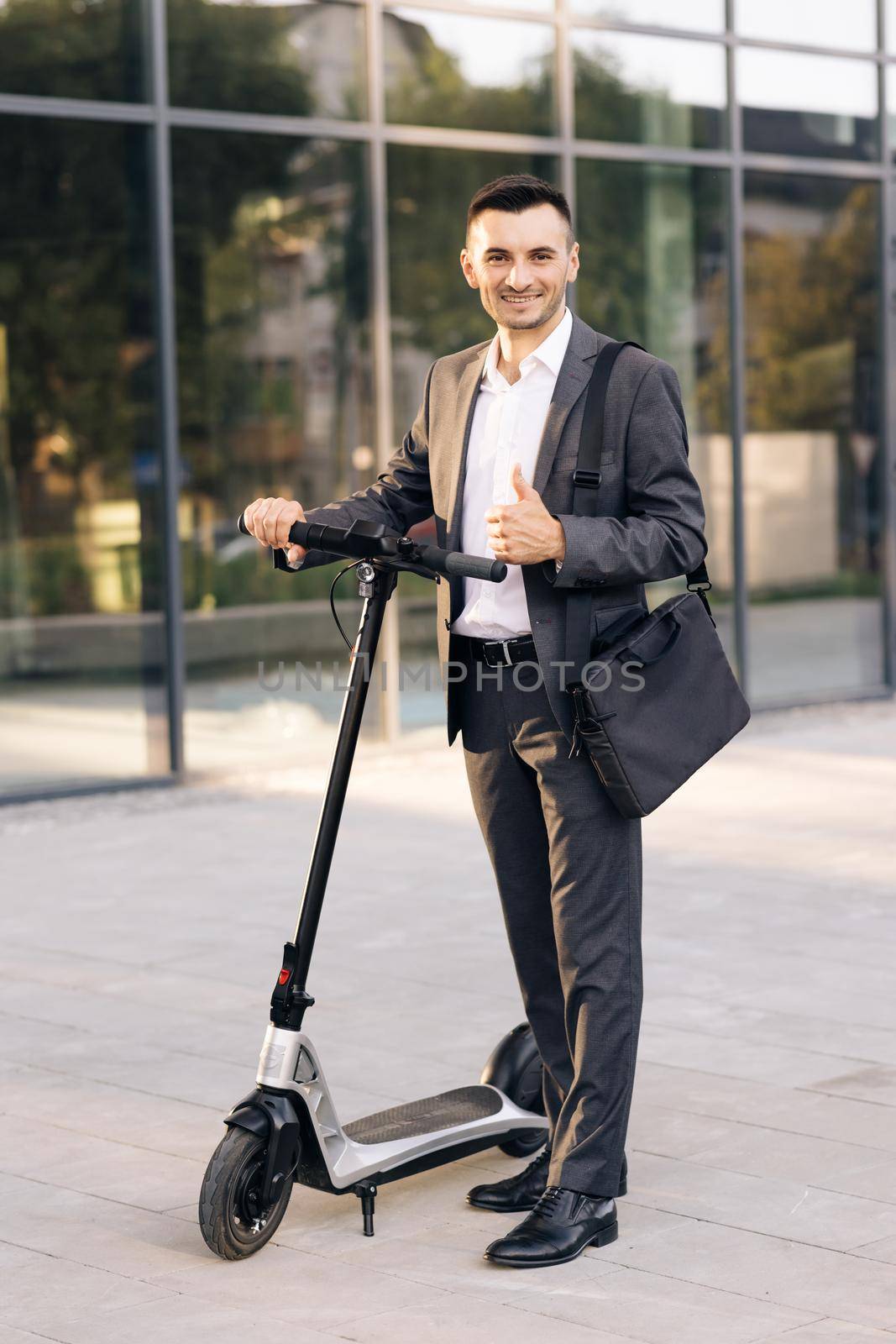 Handsome businessman with his electric scooter in front the business building. Young businessman in elegant suit standing outdoors, cheerfully smiling and showing thumb up.