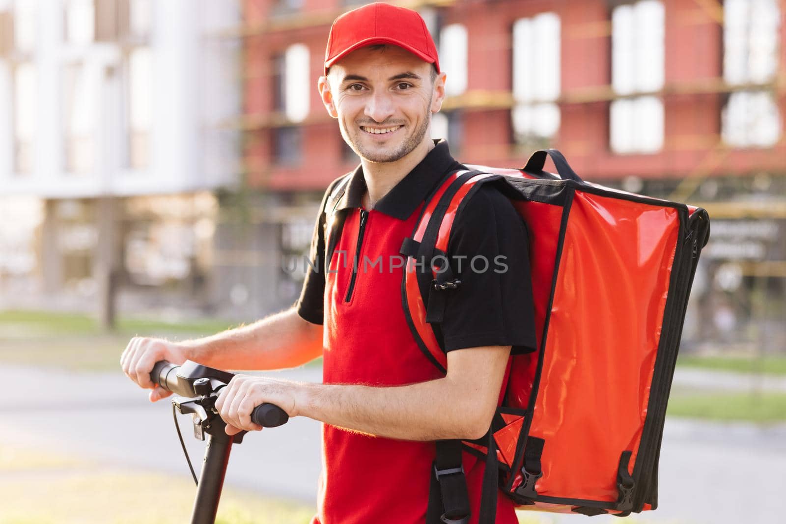 Male courier with red backpack and electric scooter. Delivering green transportation. Portrait shot of man delivery worker in red uniform standing at street and turning face to camera by uflypro