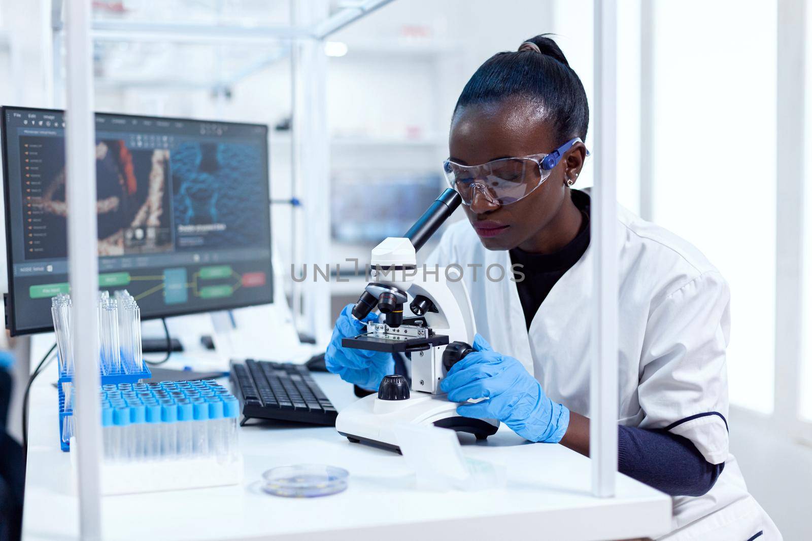 African researcher adjusting microscope lenses looking at sample on glass slide. Black healthcare scientist in biochemistry laboratory wearing sterile equipment.
