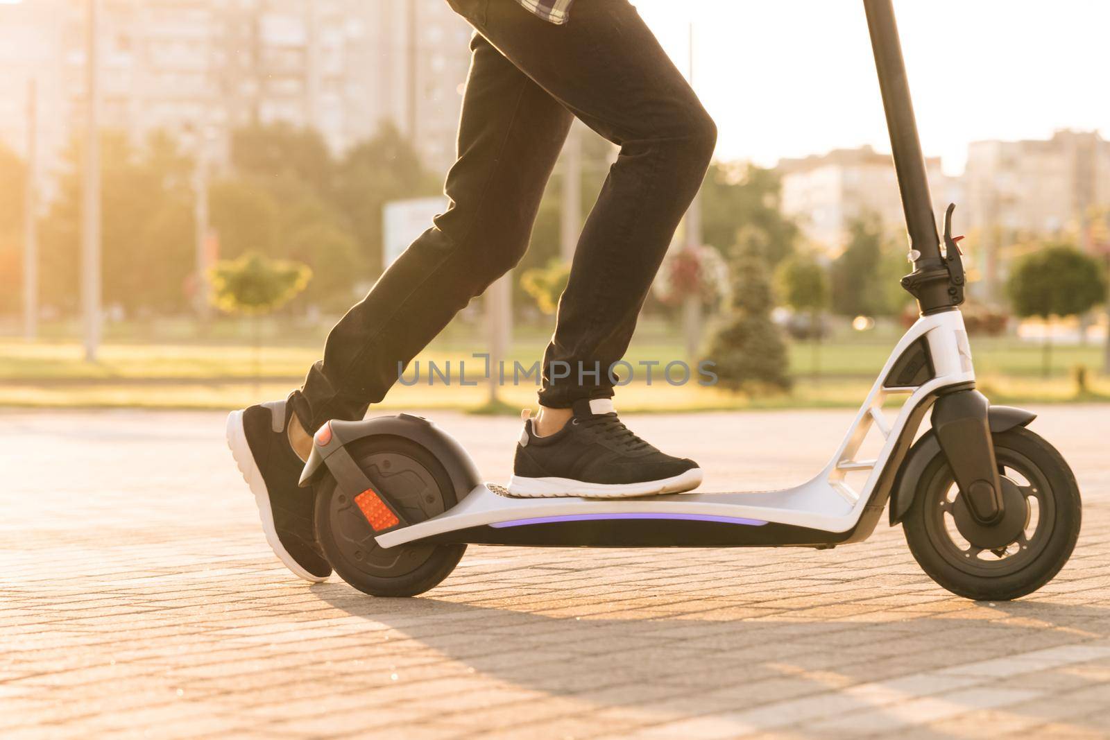 Shot on legs young man in black sneakers ride on electric mobile scooter. Close up feet man courier food delivery rides the street on electric scooter deliver online order client at sunset.