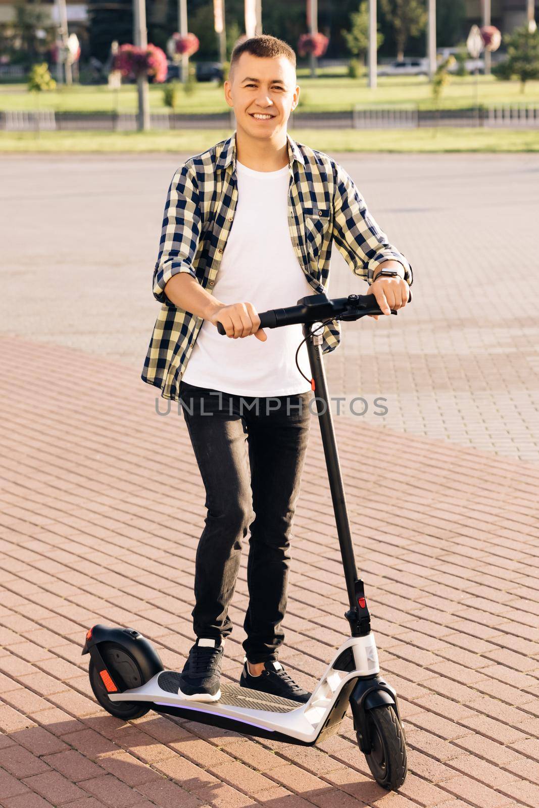 Portrait shot of caucasian hipster man leaning on electric scooter and looking at camera. Stylysh man on vehicle outdoors. Male in city in sunlight on sunset. Eco-friendly modern urban transport.