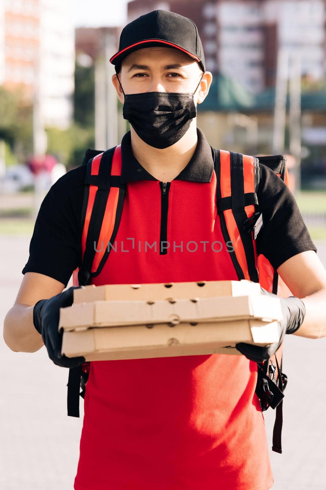 Portrait of courier delivery man with red backpack holding pizza in carton boxes, wearing protective face mask and gloves. Delivery services. Excellent delivery.