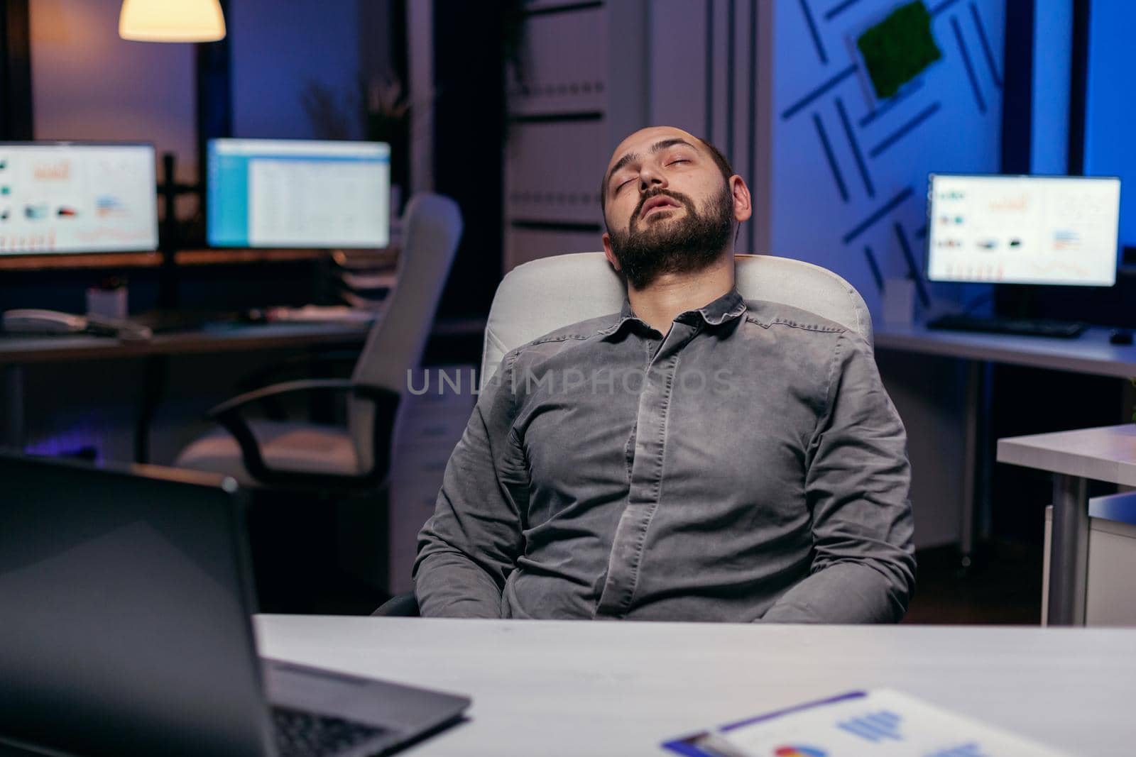 Overworked exhausted man sleeps on chair in empty office. Workaholic employee falling asleep because of while working late at night alone in the office for important company project.