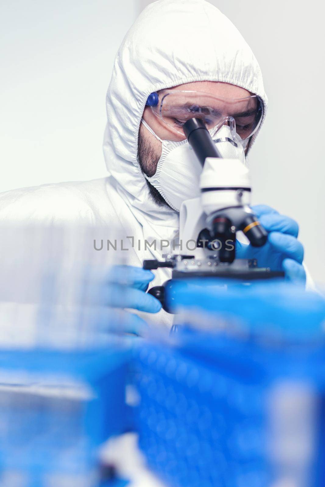 Man laborant in ppe suit using microscope doing research of coronavirus Scientist in protective suit sitting at workplace using modern medical technology during global epidemic.