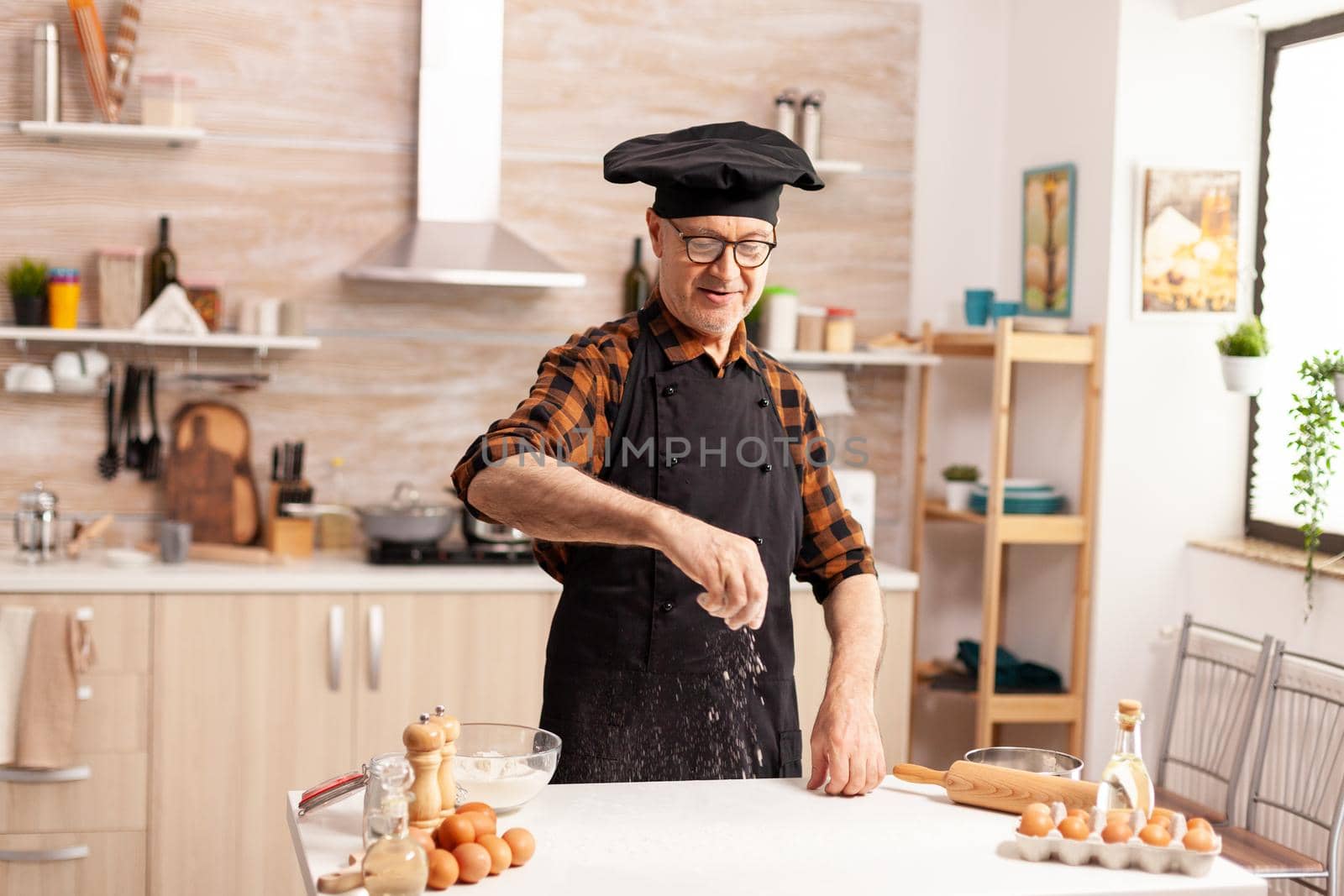Senior baker in home kitchen during preparation of cookies using wheat flour. Retired senior chef with bonete and apron, in kitchen uniform sprinkling sieving sifting ingredients by hand