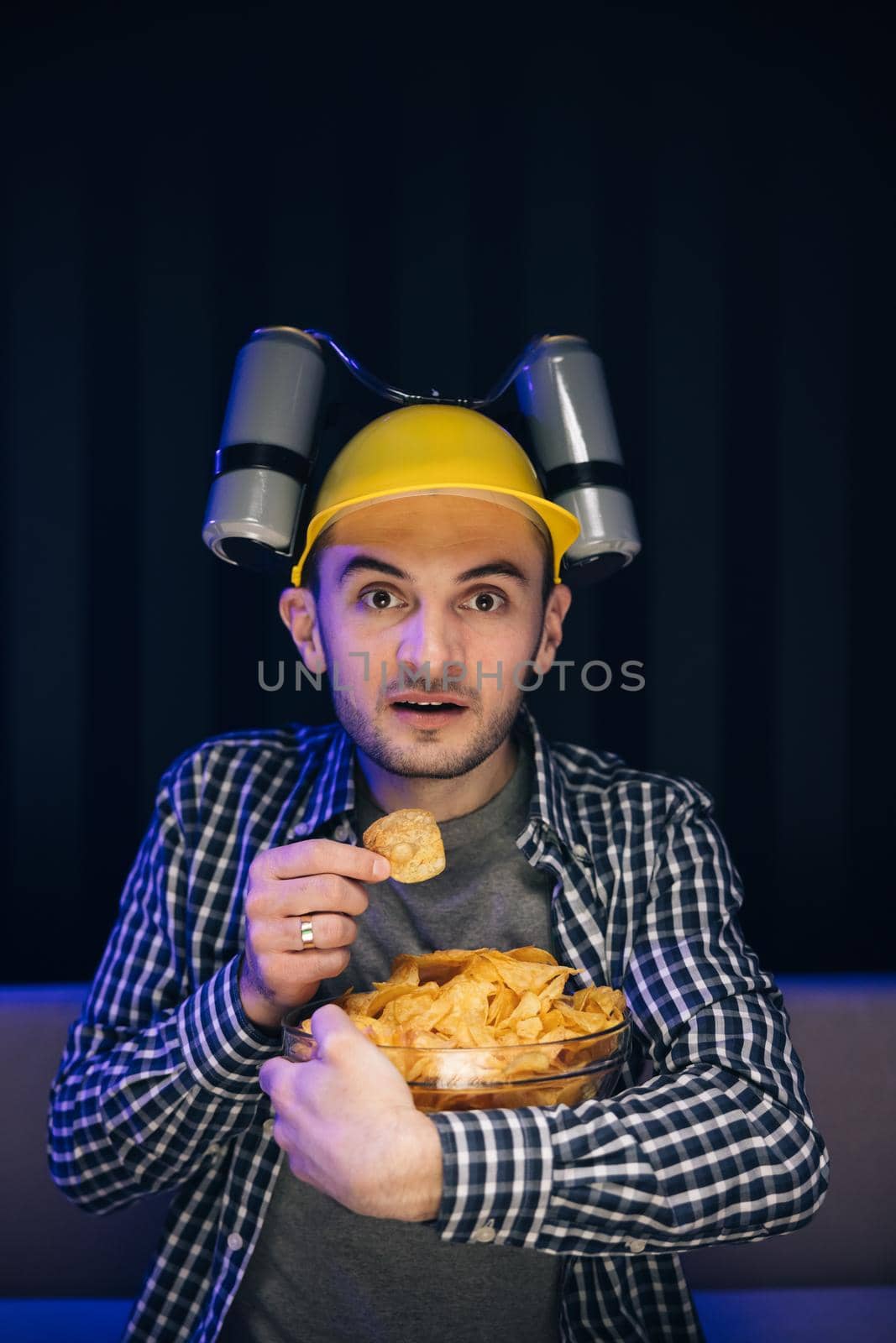 Hipster man with beer helmet on the head eating chips while watching TV At home by uflypro