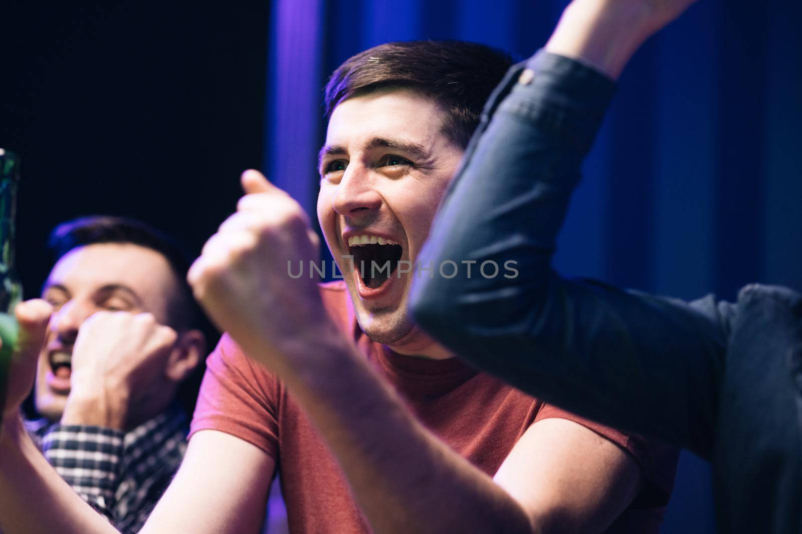 Close up of Caucasian male football fan sitting with friends at TV screen in dark living room and watching match. Cheerful man celebrating football win. Scoring goal or winning game concept.