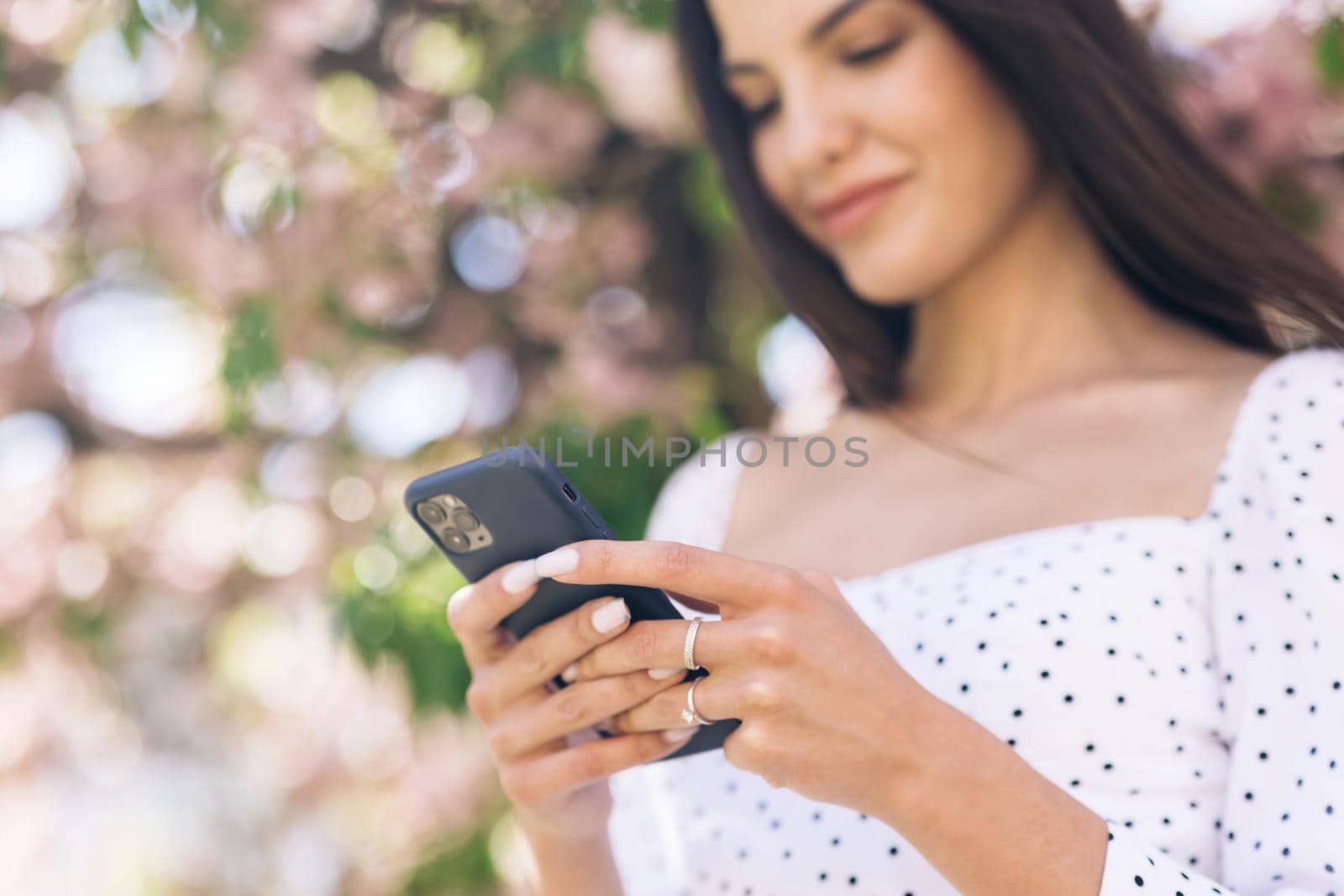Portrait of happy hipster woman typing by mobile phone outdoors. Cheerful girl walking with smartphone in urban background. Smiling lady holding cellphone in hands outside.