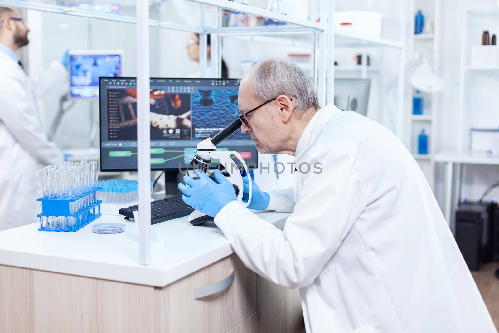 Senior scientist preparing and analyzing microscope slides with busy coworkers. Chemist researcher in sterile lab doing experiments for medical industry using modern technology.