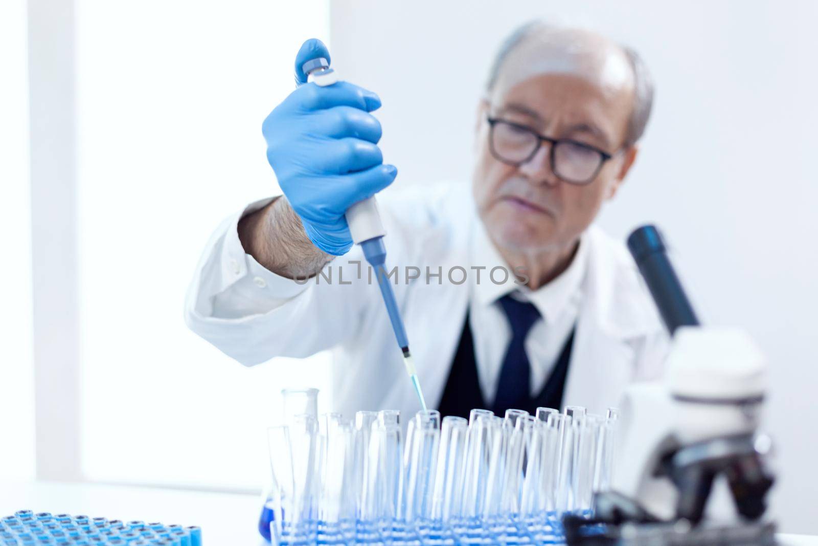 Pharmaceutical scientist pouring blue substange in test tubes sitting on his workplace with dropper pipette. Senior professional chemist using pippete with blue solution for microbiology tests.